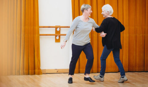 Two older women linking arms and dancing in a dance studio