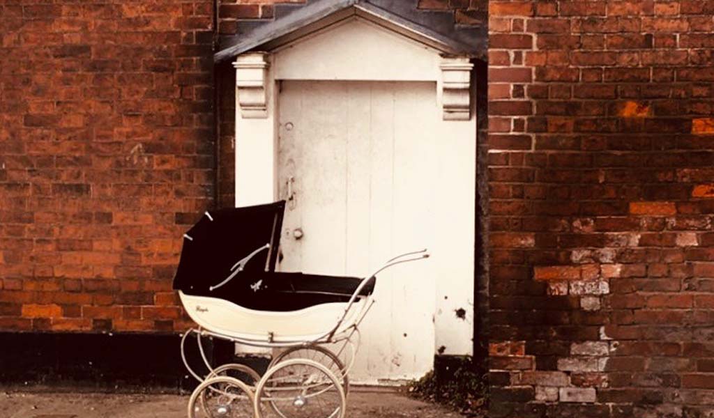 Hush Now - Old Pram station infront of a brick wall and white door