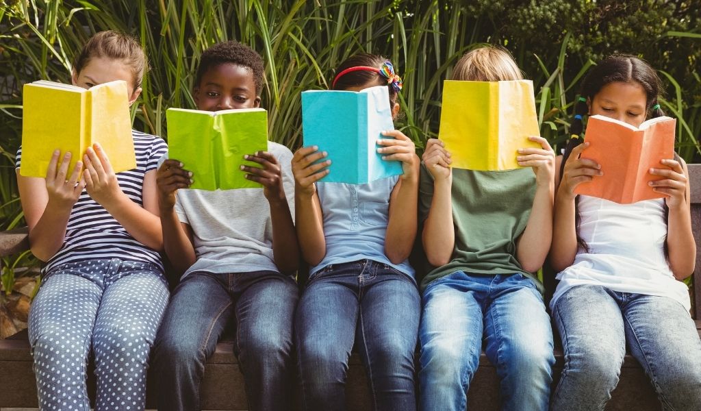 A row of children sitting on a bench, each holding a brightly coloured book up to their faces