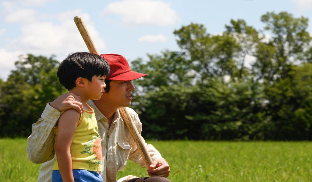 A Korean father, wearing a red cap, crouches down next to his young son with a baseball bat in his hand. Green grass and tress in the background. 