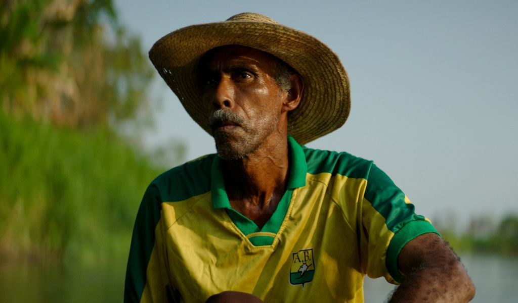 On older man in a straw hat and old yellow and green football shirt stares in to the distance in a still from the filmTantas Almas 