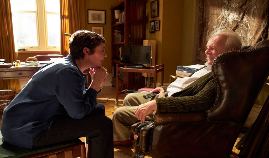 Olivia Coleman sits, smiling, with her chin resting on her hands opposite Antony Hopkins in a leather armchair - in a still from the film The Father
