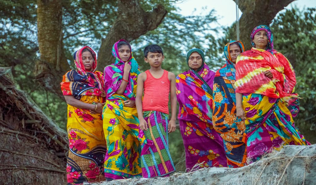 5 Bangladeshi women and a young boy stand infront of straw huts and trees wearing brightly coloured saris