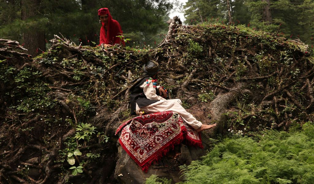 A photograph of a person wearing a turban sitting on a bright blanket in front of a tangle of exposed tree roots, whilst another person dressed in red looks on from above