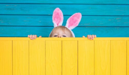 Child with Easter bunny ears on peeking over a bright yellow fence