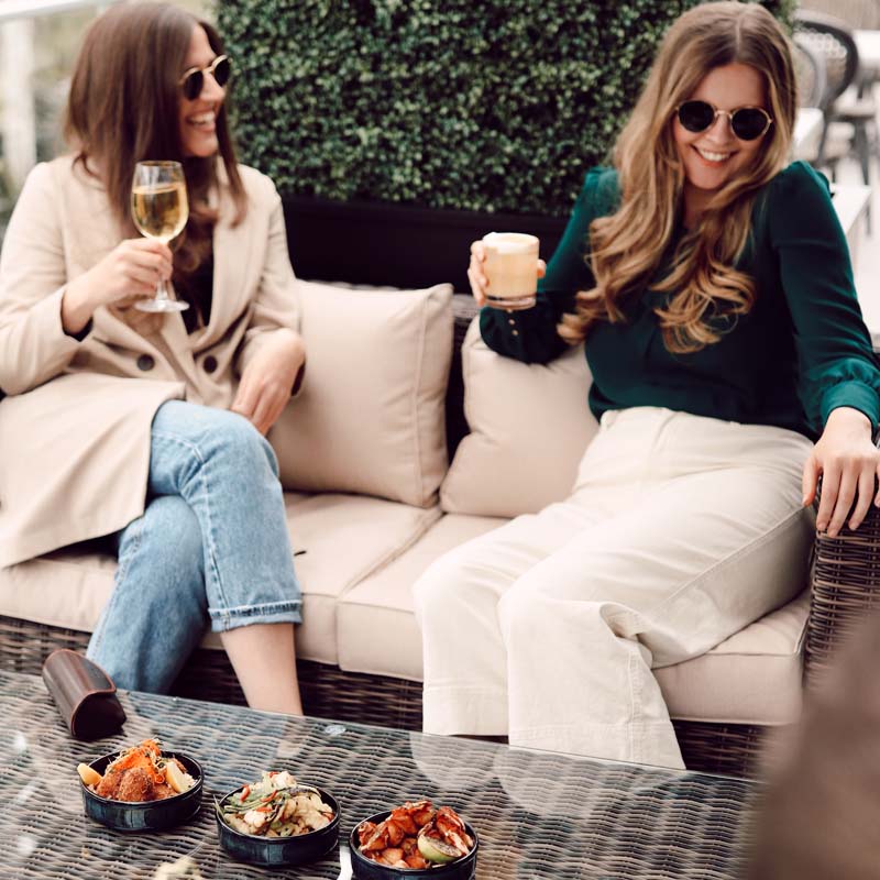Two women wearing sunglasses and holding drinks, relaxing on an outdoor sofa