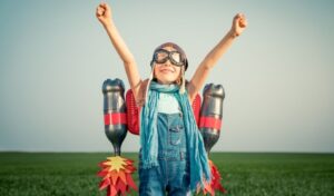 Young child wearing dungarees, a scarf and flying goggles punching the air with fists. They have a jet pack made from plastic bottles attached to them