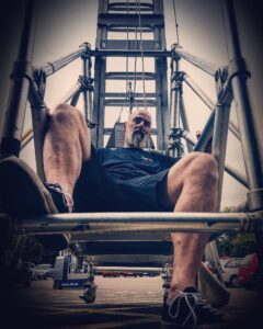 Bearded man in black shorts and tshirt, sitting on a scaffolding structure looking down at the camera