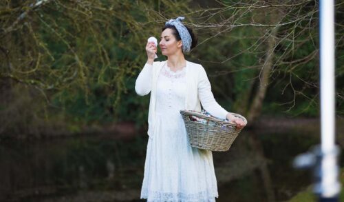 Hush Now Films image  a woman wearing a white dress and blue head scarf holds a cloth up to her nose with one hand and a basket with the other