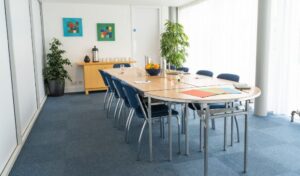 Meeting Room 1 - a long table with blue chairs either side of it, there are plants at the end of the room and colourful pictures on the wall