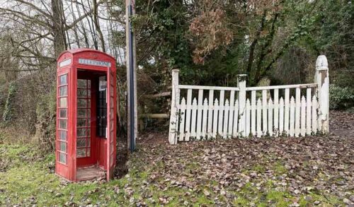 A red telephone box stands in front of trees and next to a white fence
