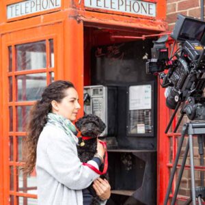 A woman holding a small black dog in her arms standing in front of a telephone box