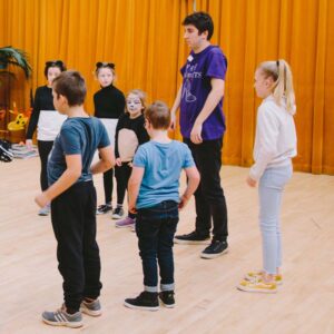 A group of children and a youth theatre leader, warming up by rolling their shoulders