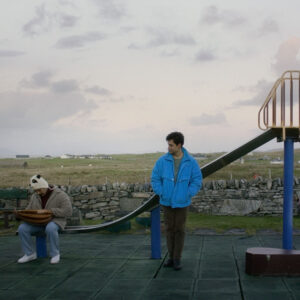 Ben Sharrock's Limbo - a man in a blue jacket stands in front of a children's slide, another man sits at the bottom of the slide looking at his lap