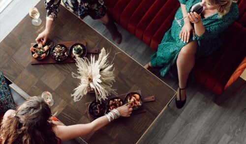 Birds eye view of women sitting on velvet sofas either side of a coffee table drinking wine and sharing a selection of tapas dishes