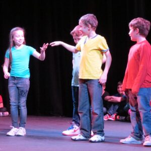 Four children in brightly coloured t-shirts, standing on stage. Two children are reaching out their hands to touch each other 