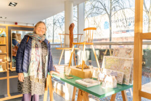 The Courtyard Shop: A lady smiling at the camera stood next to a display of wooden crafted gifts, including a chair