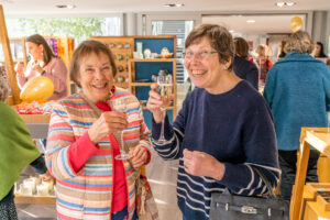Two ladies smiling at the camera holding a glass of prosecco with people browsing shop items in the background