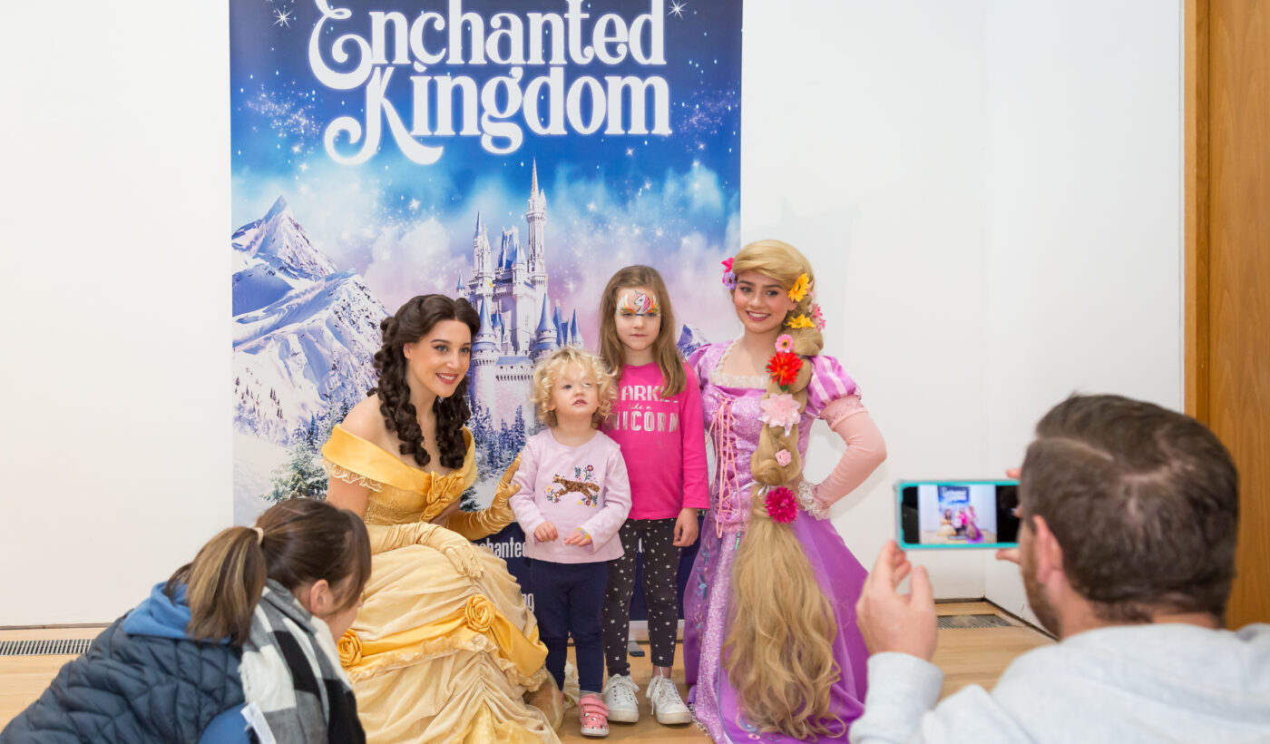 Two little girls posing for a photo with two women dressed as princesses in front of an Enchanted Kingdom sign