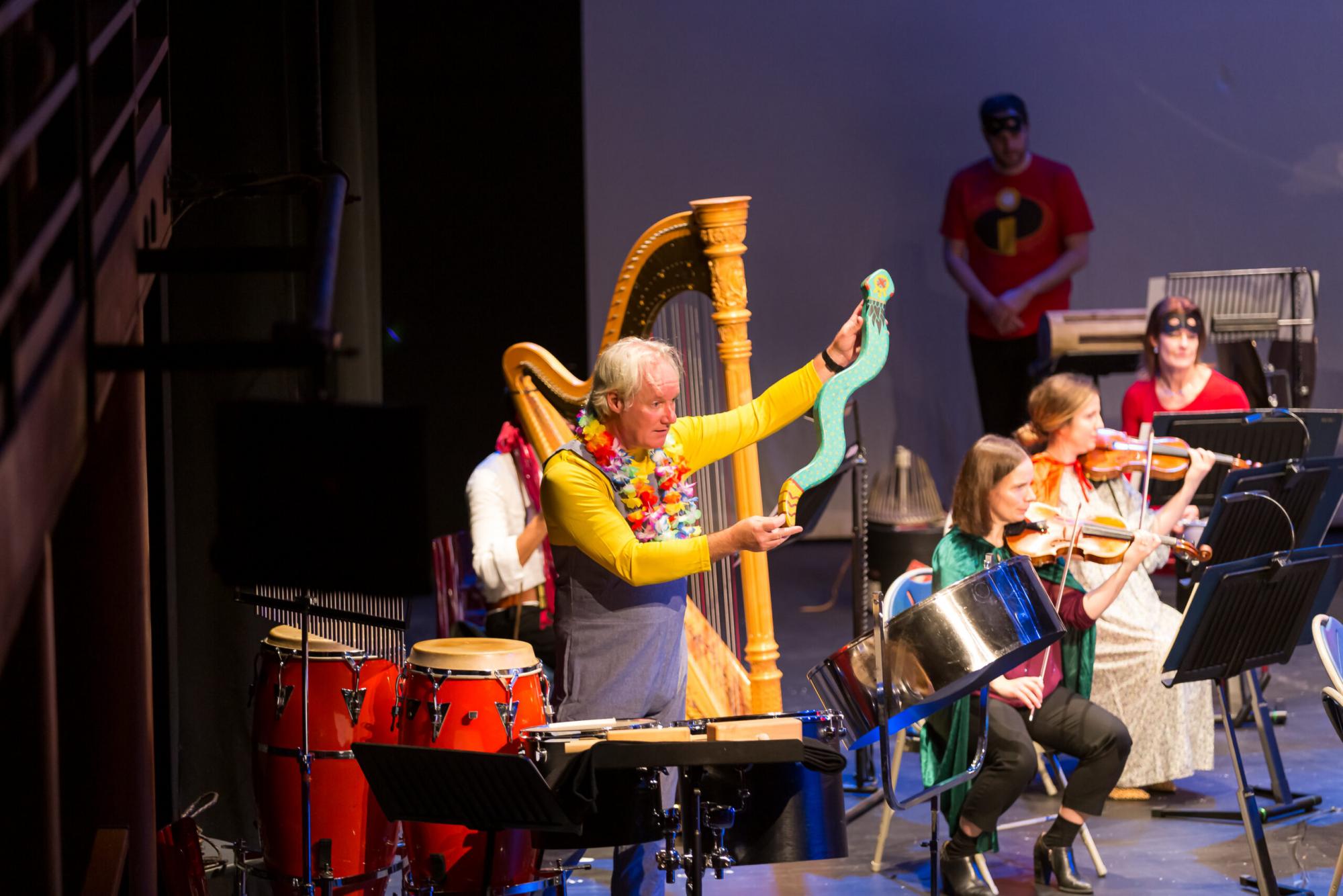 Orchestra of the Swan: A man in a minion outfit onstage holding a wooden snake