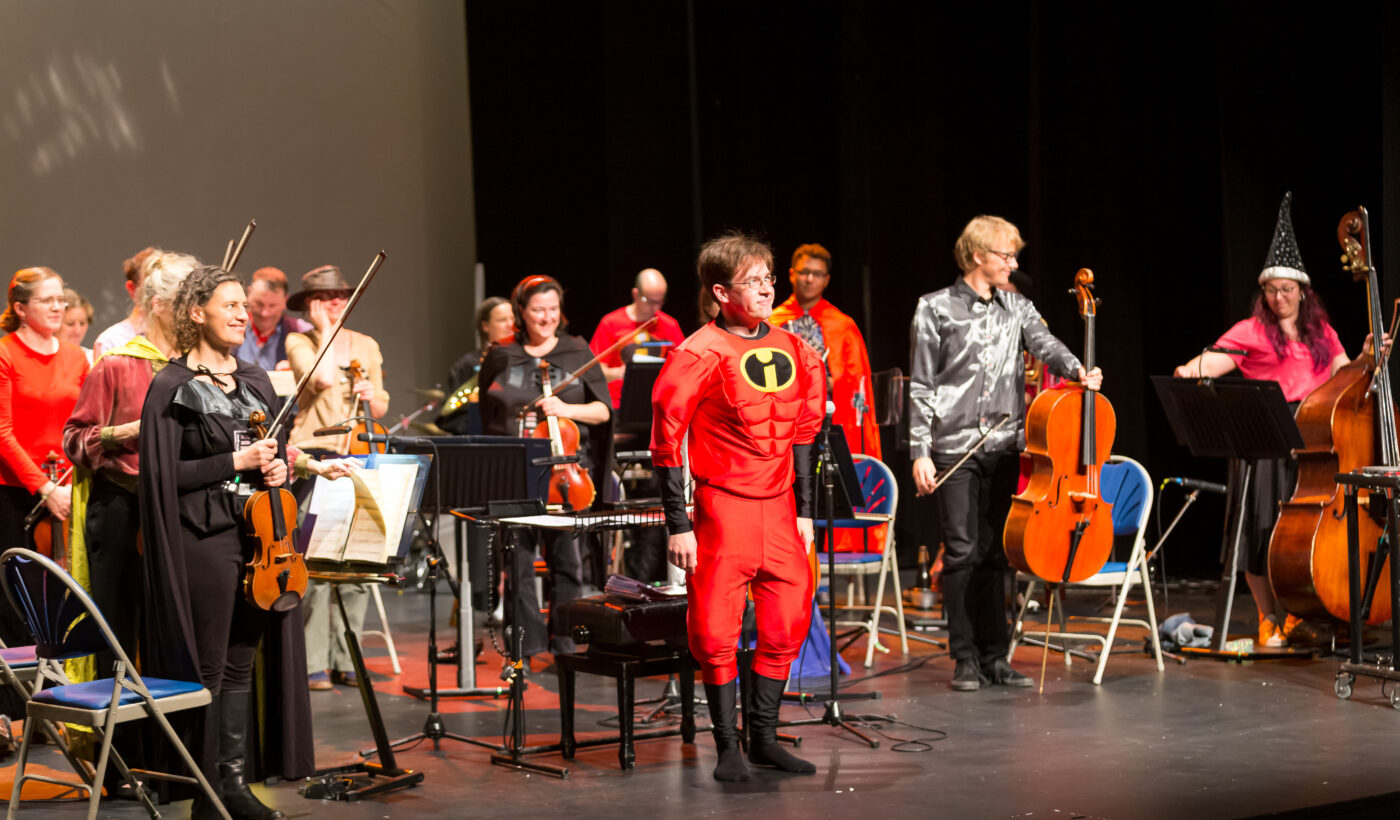 Orchestra of the Swan - an orchestra and conductor stood on stage facing the audience and holding their instruments while in fancy dress