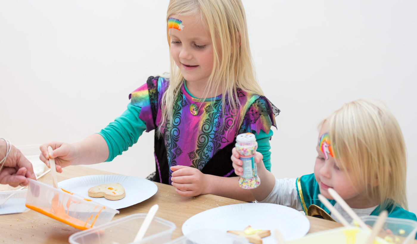 Two young girls in fancy dress stood in front of a table decorating biscuits with icing
