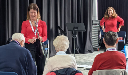 Three older people sit in a line with their backs to the camera two people are stood infront playing instruments and laughing