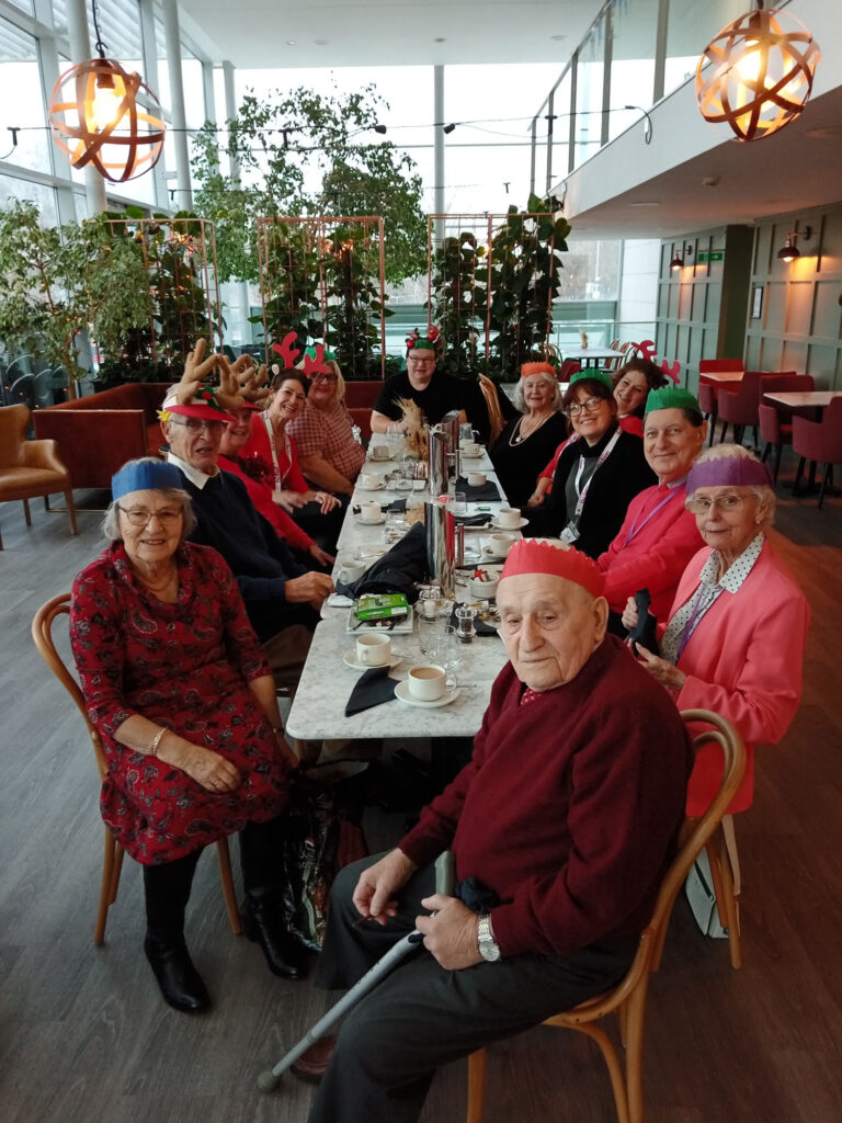 12 people sat on a long table in The Courtyard Restaurant wearing Christmas part hats and reindeer ears smiling at the camera