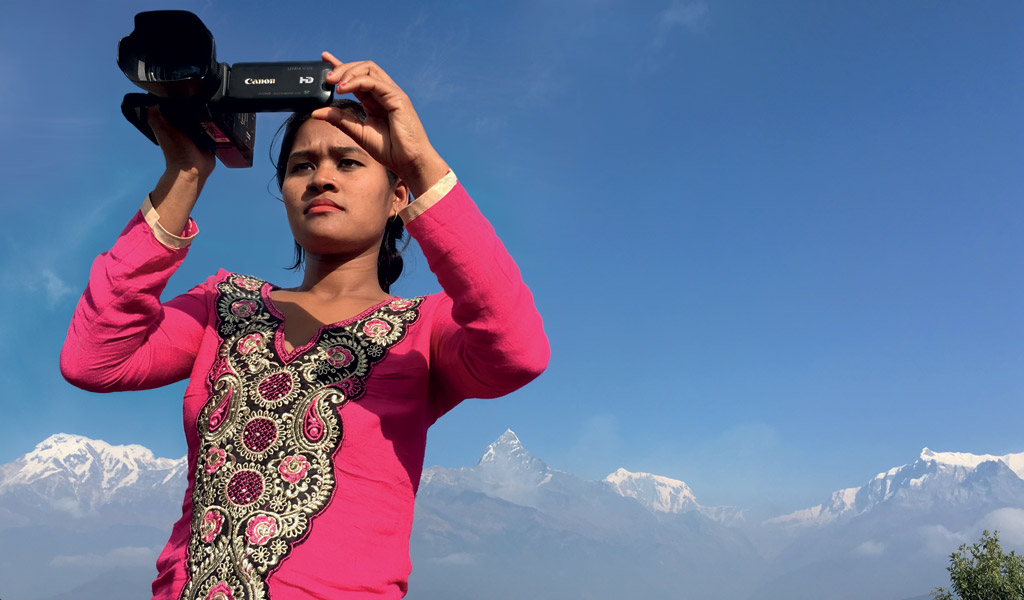 A woman in a pink dress holding up a video camera with blue skies in the background