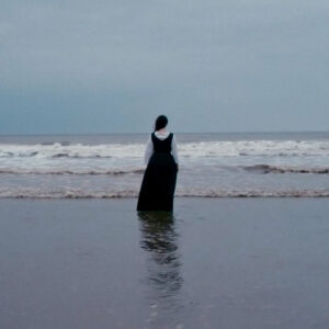 A woman in a full length black dress over a white shirt is walking towards the sea