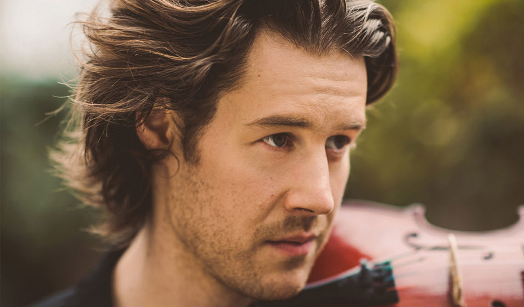 A close up of Thomas Gould looking thoughtfully into the distance with a violin under his chin