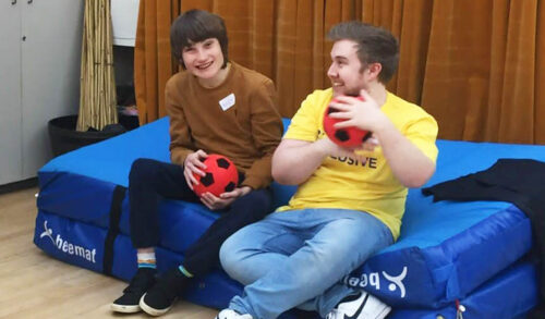 Two young men sitting on a crash mat holding footballs and smiling