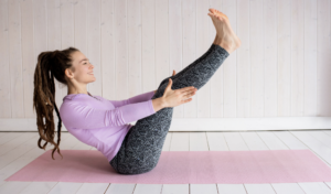 A girl in gym clothing sits on a pilates mat, holding her feet up in the air in boat pose