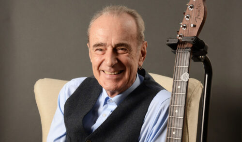 Francis Rossi with his hair in a ponytail wearing an earring and a waistcoat over a blue shirt has a guitar next to him and is smiling at the camera