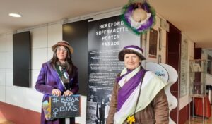 International Women's Day 2022 Two women dressed as suffragettes stood in front of a banner smiling
