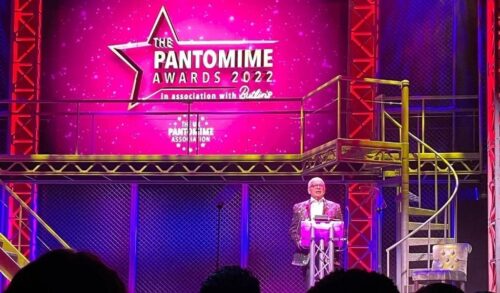 Great Bitish Panto Awards 2022  Christopher Biggins presenting onstage in front of a bright pink screen with the Pantomime Awards logo on it
