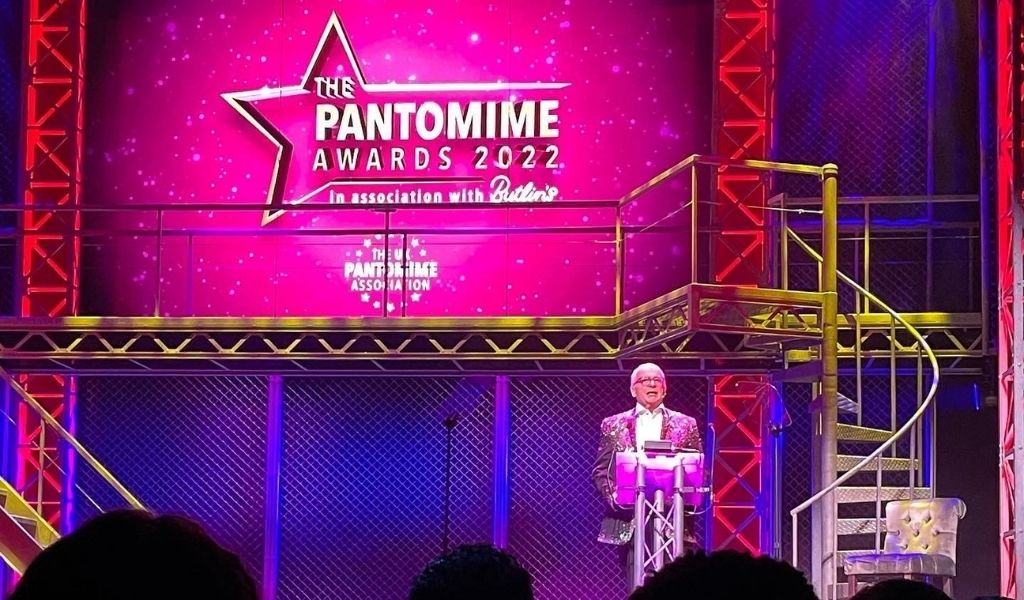 Great Bitish Panto Awards 2022 - Christopher Biggins presenting onstage in front of a bright pink screen with the Pantomime Awards logo on it