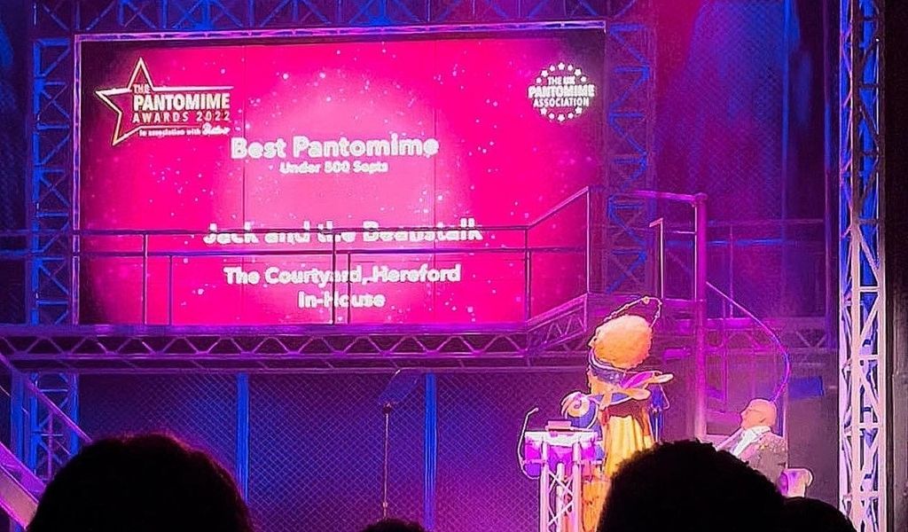 Great British Pantomime Awards 2022 - a screen with 'Best Pantomime' written on with The Courtyard listed underneath and a presenter stood in front looking at the screen.