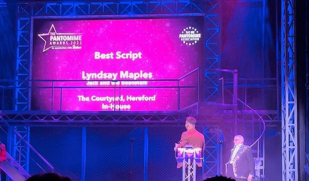 Great British Pantomime Awards 2022 - a screen with 'Best Script' written on with Lyndsay Maples listed underneath and a presenter stood in front looking at the screen.
