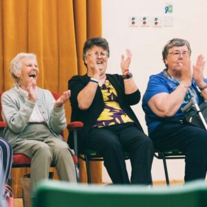 Three older ladies sitting on chairs, clapping and smiling