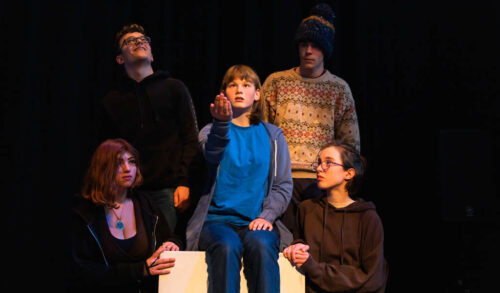 Inspired Youth Theatre Festival image a girl sitting holding her hand out surrounded by four others looking at her under a spotlight