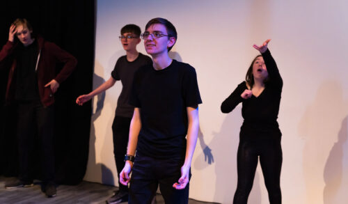 A young male actor wearing black clothes stood smiling behind him are three other performers wearing black clothes all with surprised expressions on their faces