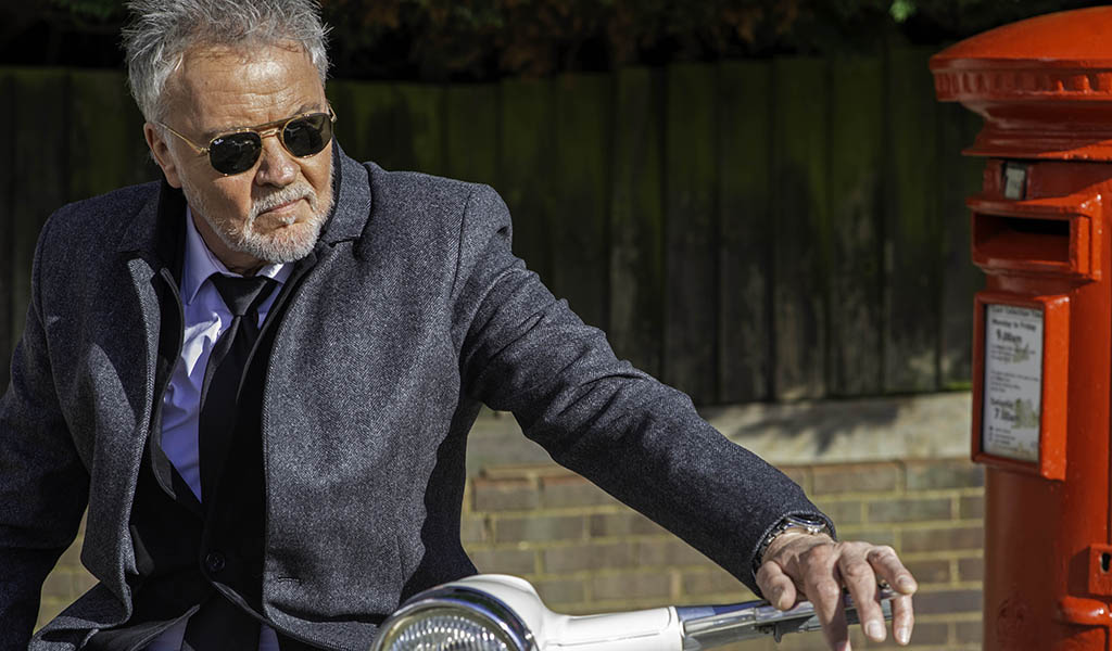 Paul Young: A man in his 60s wearing sunglasses and a suit, with white hair and beard sat on a vesper next to a red letterbox.