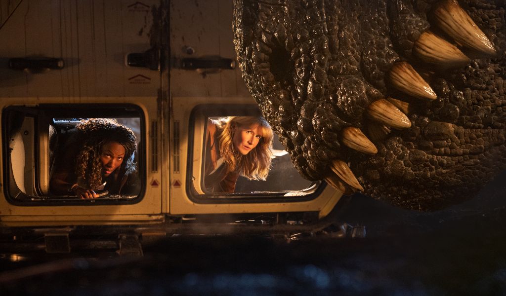 Jurassic World: Dominion (12A), Image - Two women lay in an upturned car. In front of the car is the mouth of a T-Rex