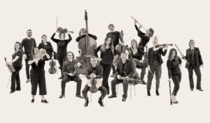 Orchestra of the Swan: A large group of musicians pose with their instruments against a cream backdrop