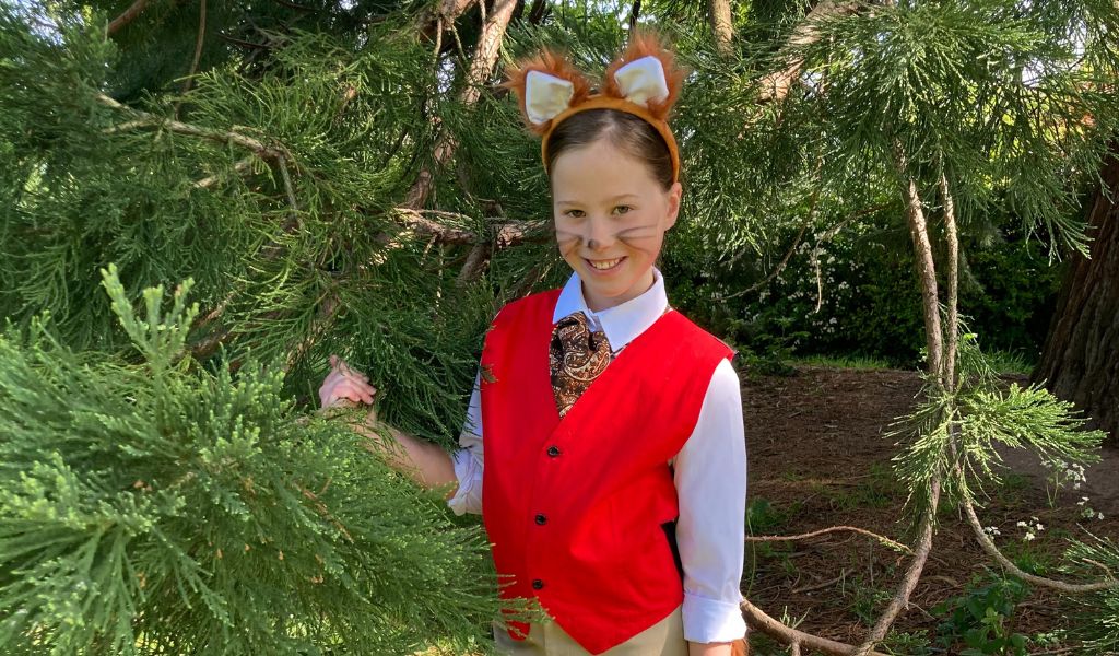 A young girl dressed in a red waistcoat wearing fox ears standing next to a tree