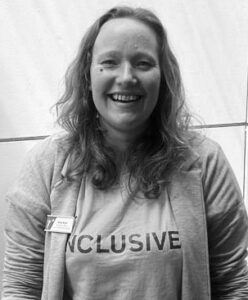 Black and white photo of Kate Ross, a woman with shoulder length hair wearing a tshirt that said 'inclusive'