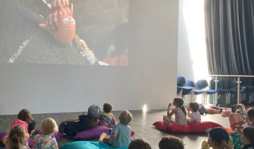 Holiday workshops image a group of children sitting on cushions watching The Snail and the Whale on a projector screen