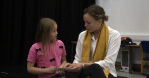 A young woman sits with a young girl, pointing at a piece of paper in her hands