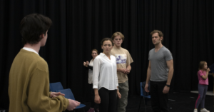 A man stands with his back to camera. A woman and two other men stand looking at him, shocked.
